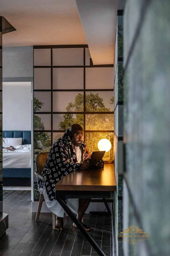 Wider interior portrait of Gabriel seated at a desk using a tablet beside a glowing lamp.