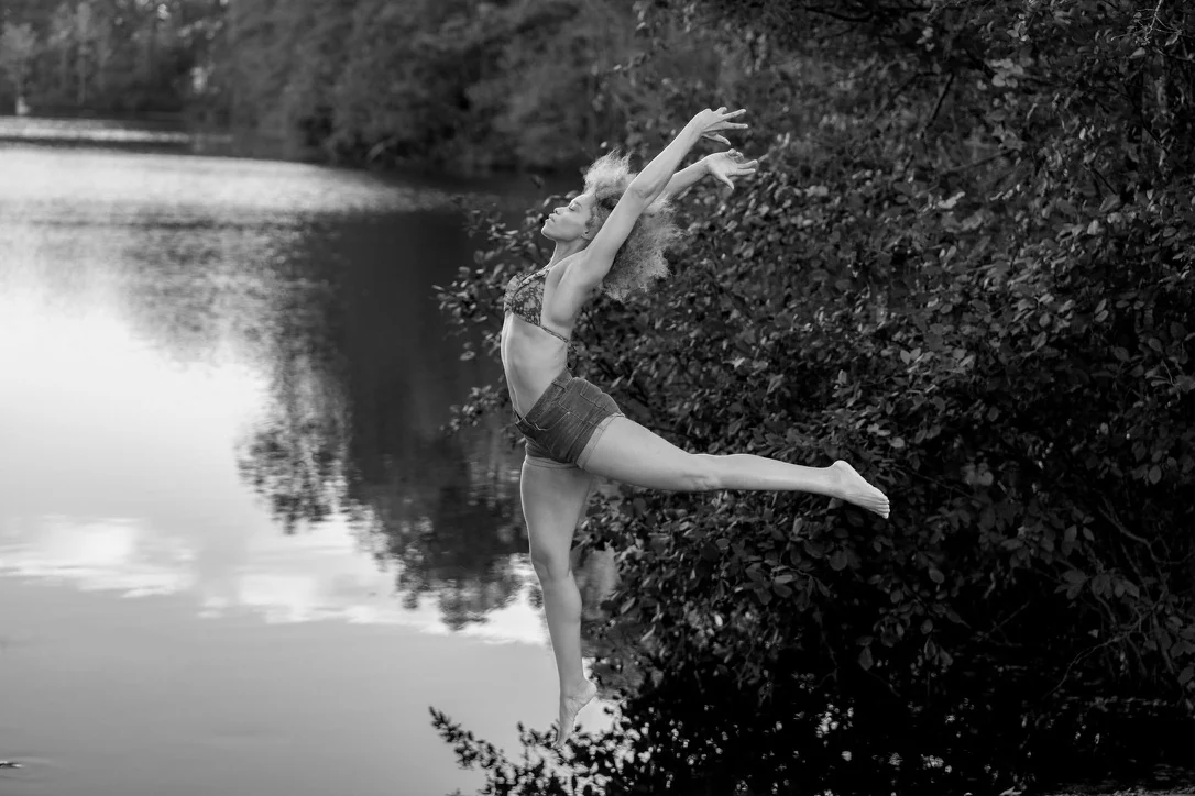 Black and white photograph of a dancer extending her arms and leg beside a lake